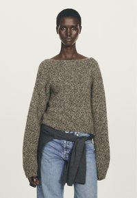 Donna che indossa un maglione a maglia marrone con maniche lunghe, jeans azzurri chiari e una felpa grigio scuro annodata in vita.