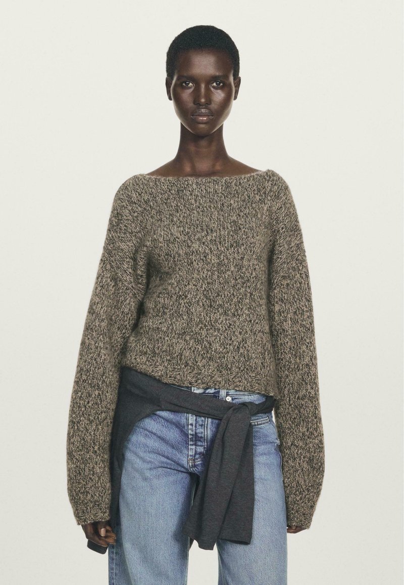 Donna che indossa un maglione a maglia marrone con maniche lunghe, jeans azzurri chiari e una felpa grigio scuro annodata in vita.