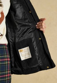 Melna jaka iekšpuse ar satīna līdzīgu tekstūru, ar etiķeti ar zelta akcentiem un sānu kabatu. Roka tur audumu.