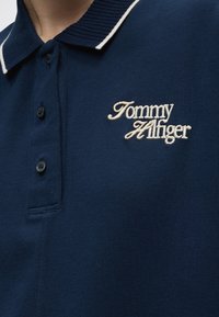 Polo bleu marine avec col bordé de blanc et texte brodé blanc "Tommy Hilfiger" sur la poitrine, trois boutons sur la patte de boutonnage.