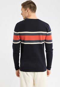 Un homme portant un pullover bleu marine avec des rayures horizontales rouges, grises et blanches sur le dos, et un pantalon blanc, tourné le dos à la caméra.