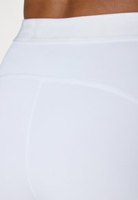 Bele športne legice bele barve z visokim pasom, ki so izdelane iz gladkega materiala, imajo ravne šive in subtilen logotip ob robu pasu.