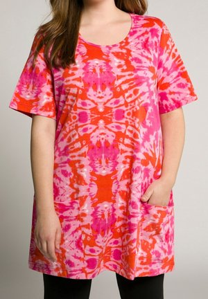 Vrouw die een losse, kortemouwige roze en oranje tie-dye tuniek draagt met twee voorzakken, staand met één hand in een zak.