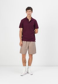 Burgundy polo skjorte, korte beige shorts, hvide sneakers, stående i et neutralt rum. Glat stof, klassisk krave, afslappet pasform.