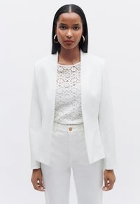 Blazer bianco su misura sopra una blouse di pizzo, abbinato a pantaloni bianchi. Tessuto morbido con design minimalista e tessuto leggero.