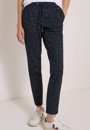 Personne portant un pantalon de jogging bleu foncé à imprimé léopard avec une taille à cordon et des baskets blanches avec des touches rouges et bleues.