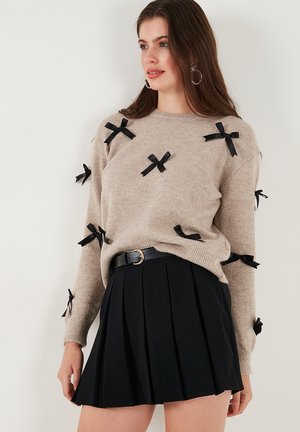 Beige knit sweater with black ribbon accents, crew neck, and long sleeves paired with a black pleated skirt and a belted waist.