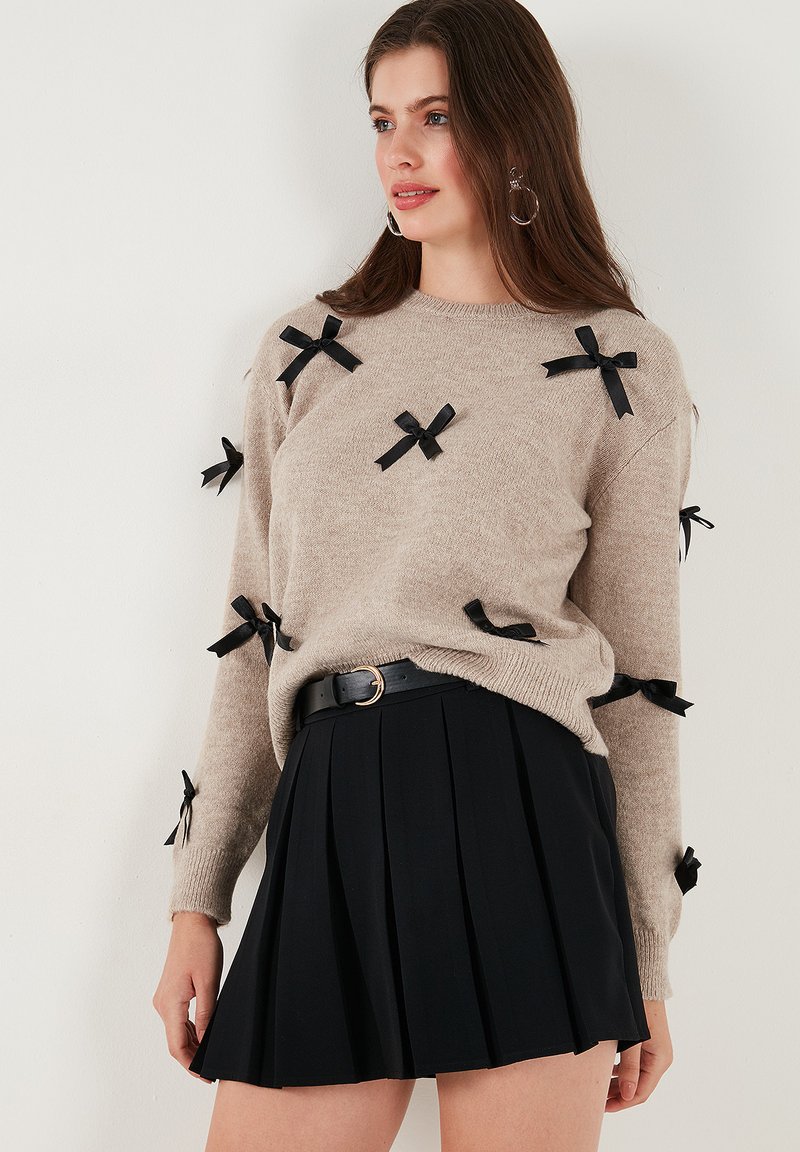 Pull en tricot beige avec des accents de ruban noir, col ras du cou et manches longues, associé à une jupe plissée noire et une taille ceinturée.