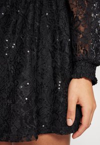 Svart spetsklänning med blommönster och paljetter, i strukturerad tyg med samlade ärmslut. Hand visas vid fållen.