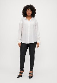 Camicia bianca con bottoni, maniche lunghe e dettagli a cravatta ai polsini, abbinata a leggings neri aderenti e scarpe con tacco alto.