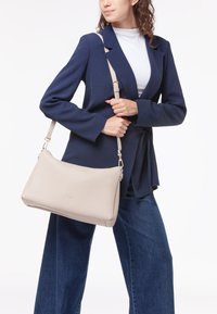 Borsa a spalla in pelle beige con una texture liscia, caratterizzata da un logo dorato e una tracolla regolabile, indossata da un modello in un blazer blu navy.