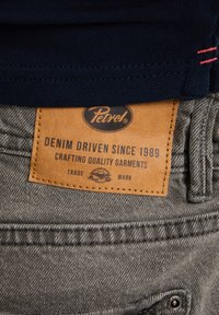 Spijkerbroek met een grijze textuur, voorzien van een beige leren label met de tekst "Petrol Denim Driven Since 1989" dat op de tailleband is gestikt.