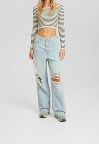 Vaqueros desgastados de color azul claro con grandes desgastes y dobladillos deshilachados, combinados con una blusa ajustada de rayas crema, sin mangas y de largo hasta los brazos.