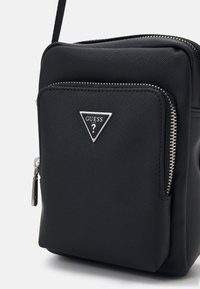 Bolso bandolera sintético negro con un logo triangular plateado, superficie texturada, bolsillo con cremallera en la parte delantera y correa ajustable. Forma compacta y rectangular.