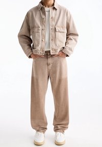 Chaqueta de mezclilla beige con dos bolsillos en el pecho y pantalones beige a juego. El modelo lleva una camiseta blanca y zapatillas blancas con acentos beige.