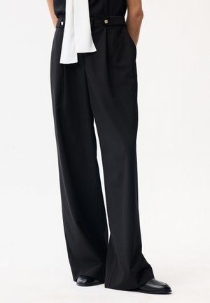 Osoba w czarnych szerokich spodniach z detalami w postaci złotych guzików, czarnych butach oraz białym dodatku materiałowym zwisającym z talii.