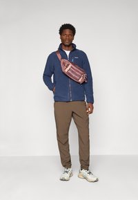 Navy fleece jacket with a zip, brown cargo pants, and a light pink sling bag featuring "Patagonia" logo, worn with gray and cream sneakers.