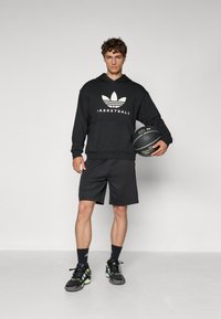Fekete kapucnis pulcsi fehér "BASKETBALL" logóval és háromlevelű dizájnnal, párosítva fekete rövidnadrággal és zöld részletekkel ellátott sportcipőkkel.