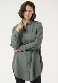 Nuppudega särk tuhmi rohelise tooniga, valmistatud tekstuurse kangaga, millel on rinnatasku ja kaardus alläär. Pika varrukaga, nööpidega mansettidega.