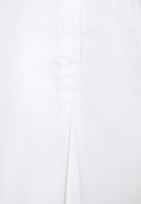 Gros plan d'un tissu blanc texturé avec une couture verticale et un détail de renfort triangulaire cousu au-dessus d'un petit pli.