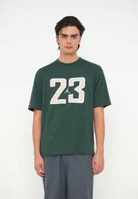 Tume roheline puuvillane T-särk ümarate kaelustega, mille esiküljel on suur heledat beeži värvi "23" trükk, lisaks väike logo aksent.