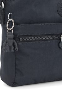 Černá nylonová taška s hladkým povrchem, obsahující kapsu na zip a malý gumový klíčenku ve tvaru opice připevněnou k tašce. Viditelný detail kruhového loga.