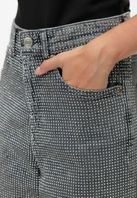Sivi traperice s srebrnim zakovicama, s prilagođenim dizajnom, prednjim džepom, kopčanjem na gumb te teksturiranom površinom za dodatne detalje.