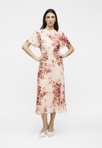 Γυναίκα με ροζ λουλουδάτο φόρεμα μήκους μέχρι τη μέση με φτερωτά μανίκια και λευκά παπούτσια με μυτερό τούβλο, στέκεται με τα χέρια στη μέση.