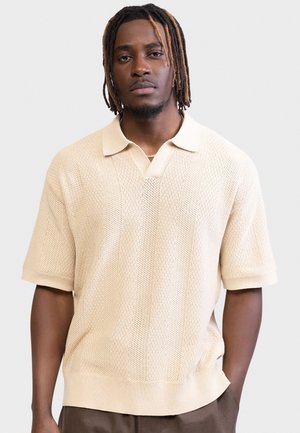 Μαύρος άνδρας με dreadlocks μέχρι τον ώμο, ντυμένος με μπεζ κοντομάνικη πλεκτή πόλο μπλούζα και καφέ παντελόνια, στέκεται μπροστά σε ένα φωτεινό φόντο.