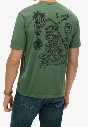 Mężczyzna w zielonej koszulce z czarnym tygrysem, bambusem, okrągłym emblem i japońskim stylem tekstu na plecach, w zestawieniu z niebieskimi dżinsami.