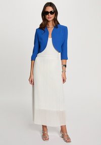Blauw cropped jasje over een witte geplooide maxi jurk. De jurk heeft verticale plooien en de outfit wordt compleet gemaakt met zilveren bandjesandalen.
