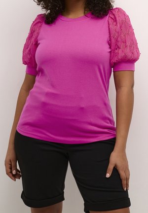 Fuksjowy t-shirt z teksturowanymi koronkowymi bufkami na rękawach, połączony z czarnymi szortami sięgającymi do kolan. Dwukolorowy wzór łączy gładkie i wzorzyste materiały.