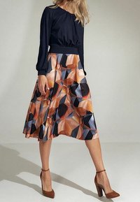 Femme portant un haut noir à manches longues et une jupe midi à motif géométrique dans des tons marron, bleu et orange, accompagnée de talons hauts marron.