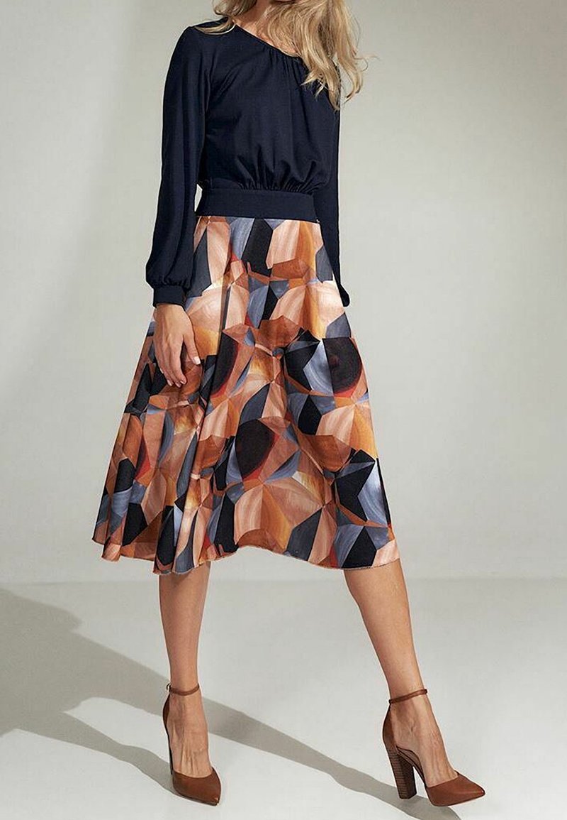 Femme portant un haut noir à manches longues et une jupe midi à motif géométrique dans des tons marron, bleu et orange, accompagnée de talons hauts marron.