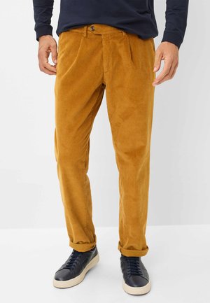 Corduroy broeken in mosterdgeel met verticale ribbels, met een riemlus, knoopsluiting en opgerolde manchetten. Gepaard met marineblauwe sneakers.