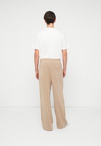 Pantaloni din bumbac bej cu o croială lejeră, talie elastică și două buzunare la spate. Textură netedă, picioare largi, purtați cu un tricou alb.