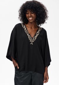 Schwarze Tunika mit einem breiten V-Ausschnitt, der mit aufwendiger beiger Stickerei verziert ist. Lockerer Schnitt mit weiten Ärmeln, aus einem weichen Stoff gefertigt.