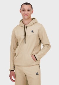 Sudadera beige con cordón ajustable, bolsillo canguro y detalles en negro. Combinada con pantalones a juego, presenta un logo en el pecho.