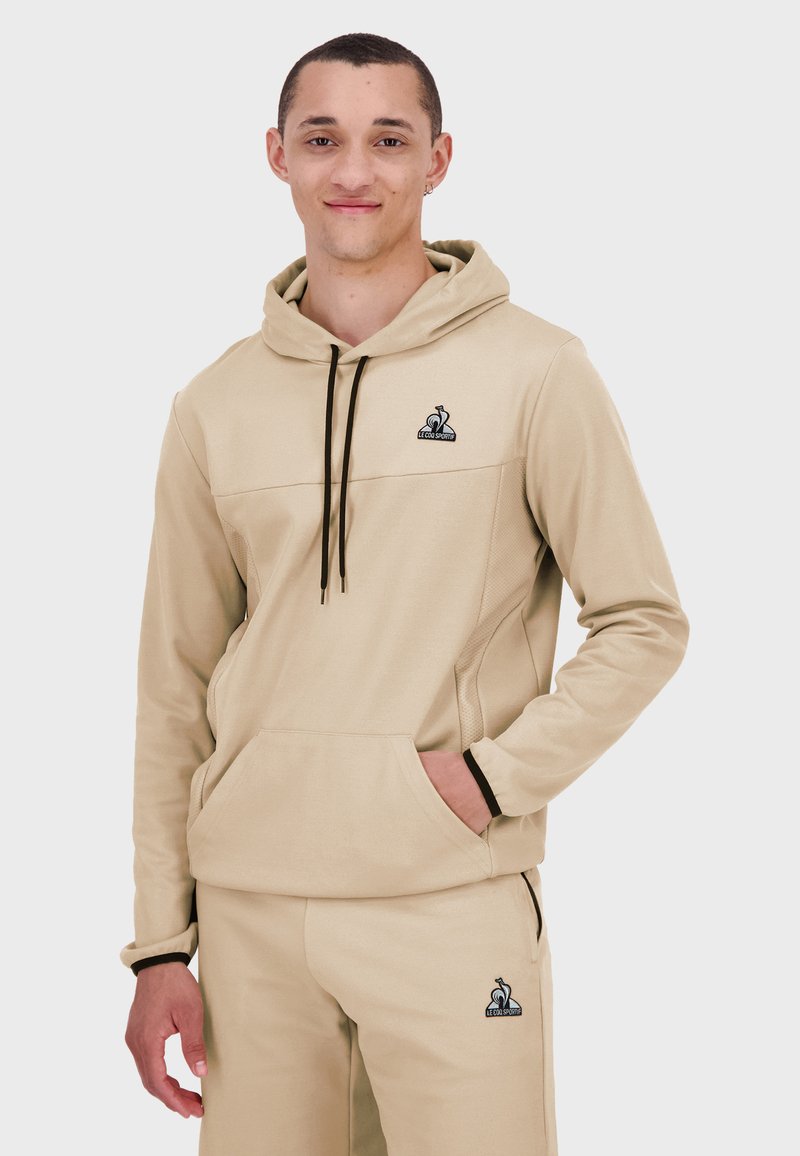 Sudadera beige con cordón ajustable, bolsillo canguro y detalles en negro. Combinada con pantalones a juego, presenta un logo en el pecho.