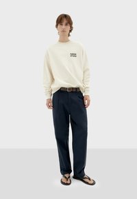 Crème sweatshirt met "FUNGHISTUDIO" in zwart gedrukt, gecombineerd met donkerblauwe plooibroek en zwarte slippers. Eenvoudig ontwerp, comfortabele pasvorm.