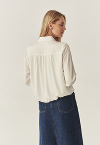 Cămașă crem cu nasturi, dotată cu un spate cu yoke și pliere, asortată cu o fustă din denim albastru închis. Material neted, cu o potrivire lejeră.