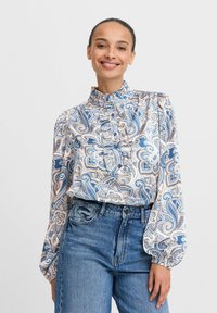 Blusa a maniche lunghe con motivo paisley, dotata di un colletto arricciato alto, in tonalità blu, arancione e beige, e chiusura frontale con bottoni. Indossata con jeans in denim.