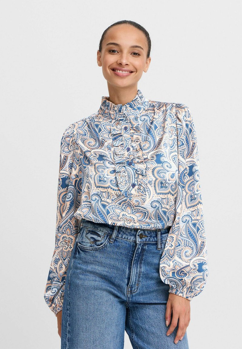 Blusa a maniche lunghe con motivo paisley, dotata di un colletto arricciato alto, in tonalità blu, arancione e beige, e chiusura frontale con bottoni. Indossata con jeans in denim.