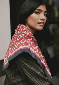 Foulard di seta con un pattern geometrico rosso e beige, bordato da un bordo blu scuro, drappeggiato elegantemente attorno al collo di una persona.