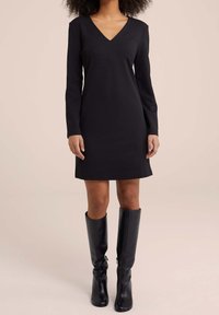 Czarna sukienka z długim rękawem o dekolcie w kształcie litery V, dopasowana w talii, o gładkiej, miękkiej fakturze; zestawiona z czarnymi kozakami do kolan.