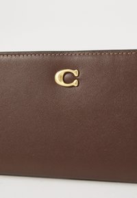 Brown leather wallet with gold metal "C" logo centered near the top edge and visible stitching along the top border.