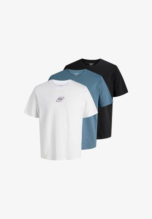 Fiatal férfi áll, fehér Jack & Jones pólót, laza sötét farmert és barna cipőt visel egy egyszínű világos háttér előtt.