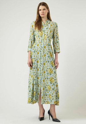 Kvinde iført en lyseblå, lang blomsterkjole med gule blomster og trekvartærmer, kombineret med sorte højhælede sko.