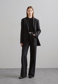 Blazer noir avec épaules structurées, associé à un haut noir ajusté à côtes et à un jean noir taille haute à jambes larges. Des chaussures noires complètent la tenue.