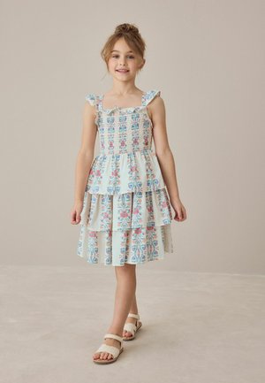 Fată tânără stând și zâmbind, purtând o rochie florală în straturi cu bretele volanate și sandale albe, pe un fundal neutru.